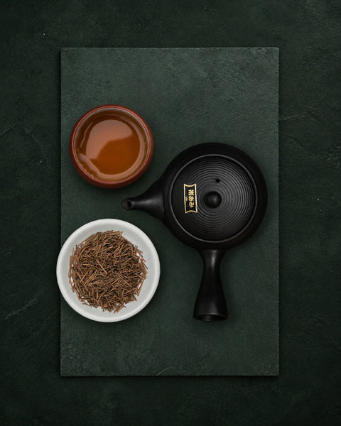 A black ceramic teapot with gold calligraphy and a small brown cup filled with hojicha tea, alongside a white bowl of kuki hojicha loose tea leaves, arranged on a dark green slate board.