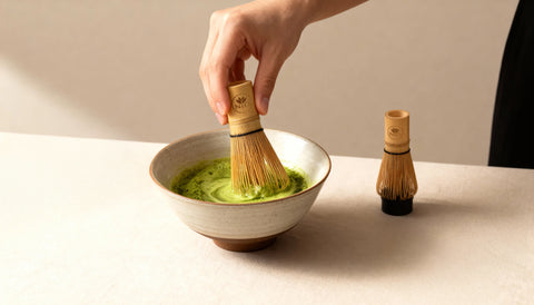 A hand holds a bamboo matcha whisk over a ceramic bowl filled with green matcha powder, with a second whisk standing on the table beside it.