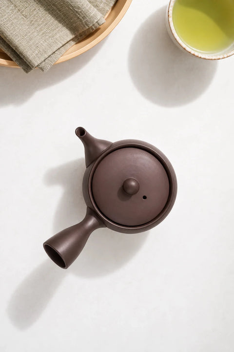 A dark brown ceramic teapot with a rounded body, lid, and protruding spout, photographed on a light beige surface with soft shadows and a few tea leaves nearby.