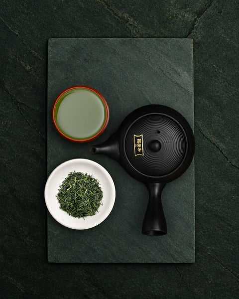 A black ceramic teapot with gold Japanese characters on the lid, arranged on a dark green slate tray with a cup of sencha shizuku green tea, loose leaf tea, and a small white dish on a dark green textured background.