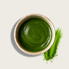 A top-down view of a small speckled ceramic bowl filled with smooth bright green matcha powder, with a matching green powder smear beside it on a light warm gray background.