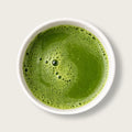 A top-down view of a vibrant green matcha tea or green juice mixture in a white ceramic bowl, set against a clean, light beige background.